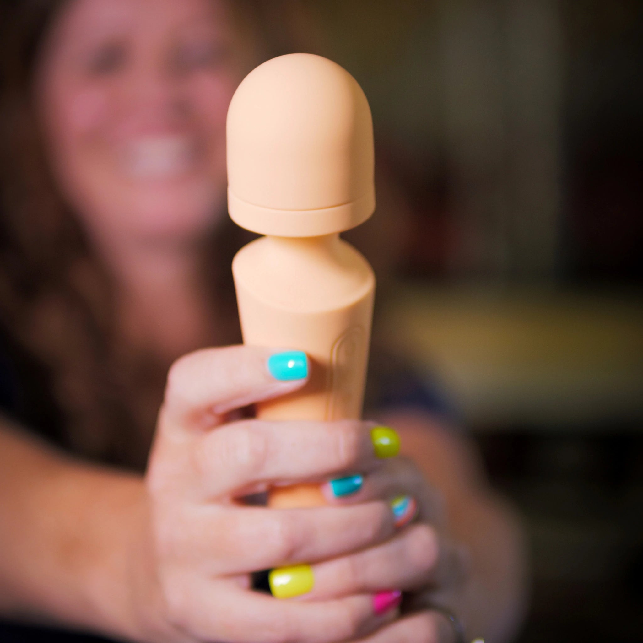 A smiling woman holding a peach-colored wand massager with bright multicolored nails in focus.