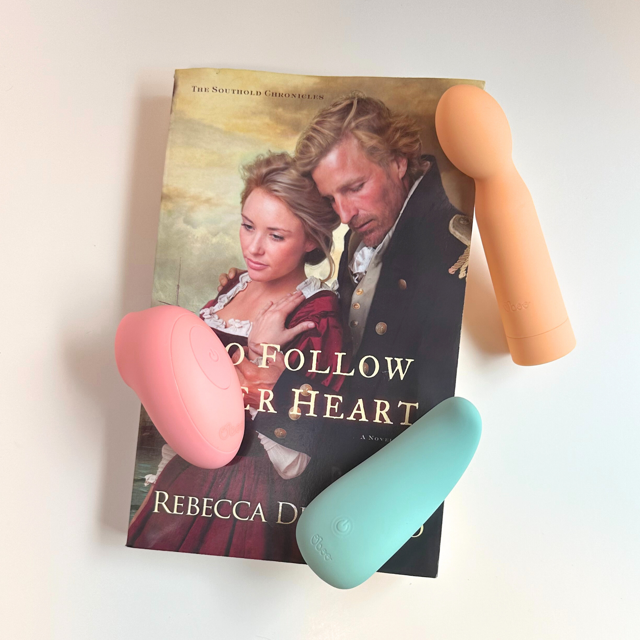 Three colorful Oboo massagers are arranged on top of a romance novel titled “To Follow Her Heart” by Rebecca DeMarino.