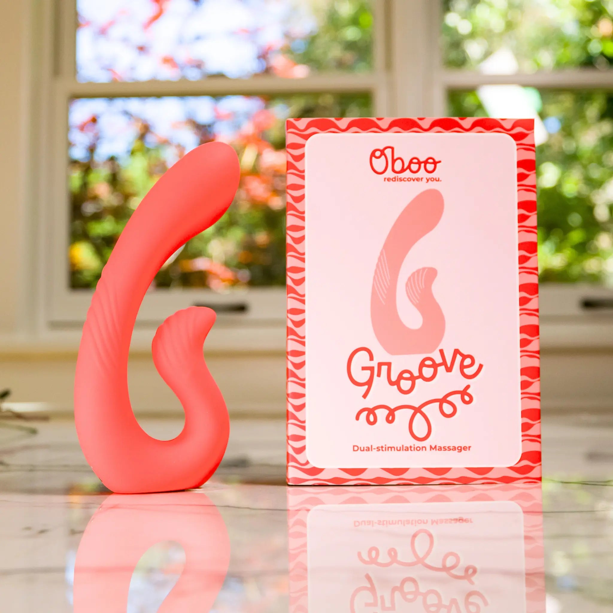 Coral Groove Dual-Stimulation Massager beside its Oboo rediscover-you product box on a marble counter with a sunny window in the background
