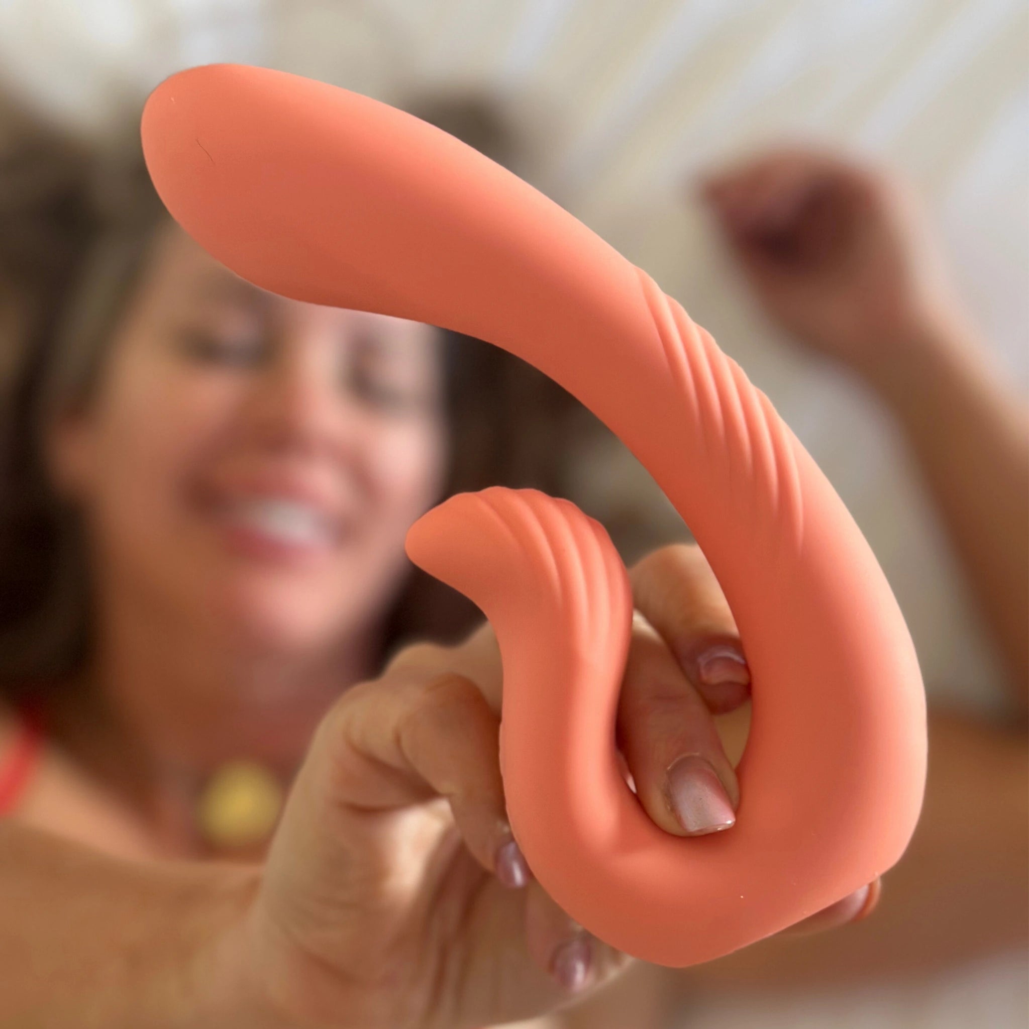 A smiling woman lying down holds a coral-colored dual-stimulation massager close to the camera, highlighting its curved, textured design.