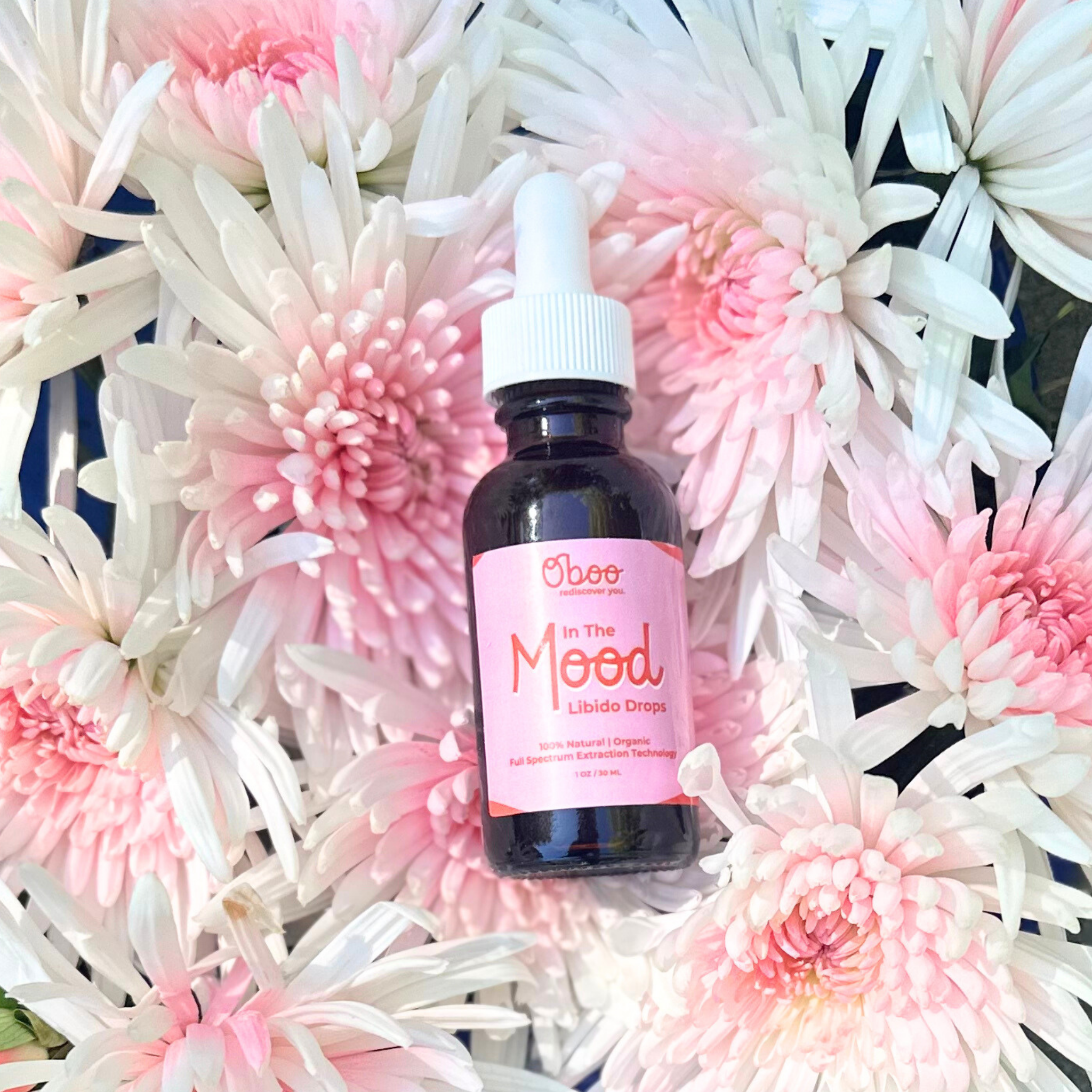 “In The Mood” libido drops bottle placed among soft pink and white chrysanthemum flowers on a natural flat lay.