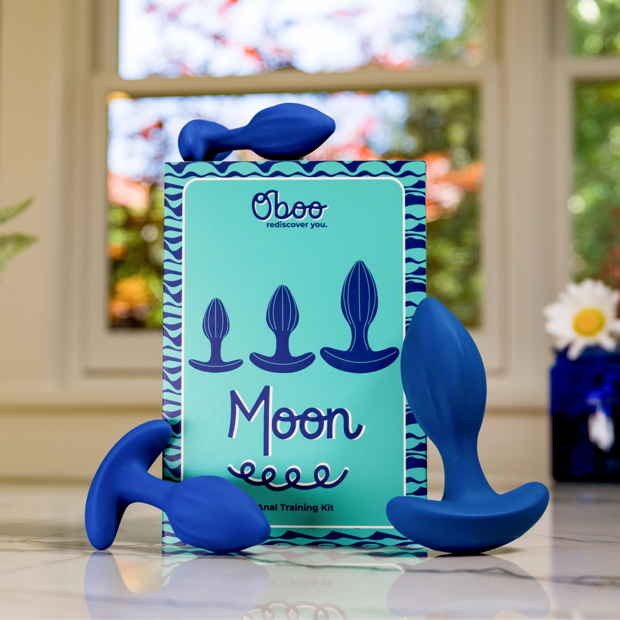 A blue anal training kit with four differently sized plugs is displayed next to the Oboo Moon product box on a reflective surface.