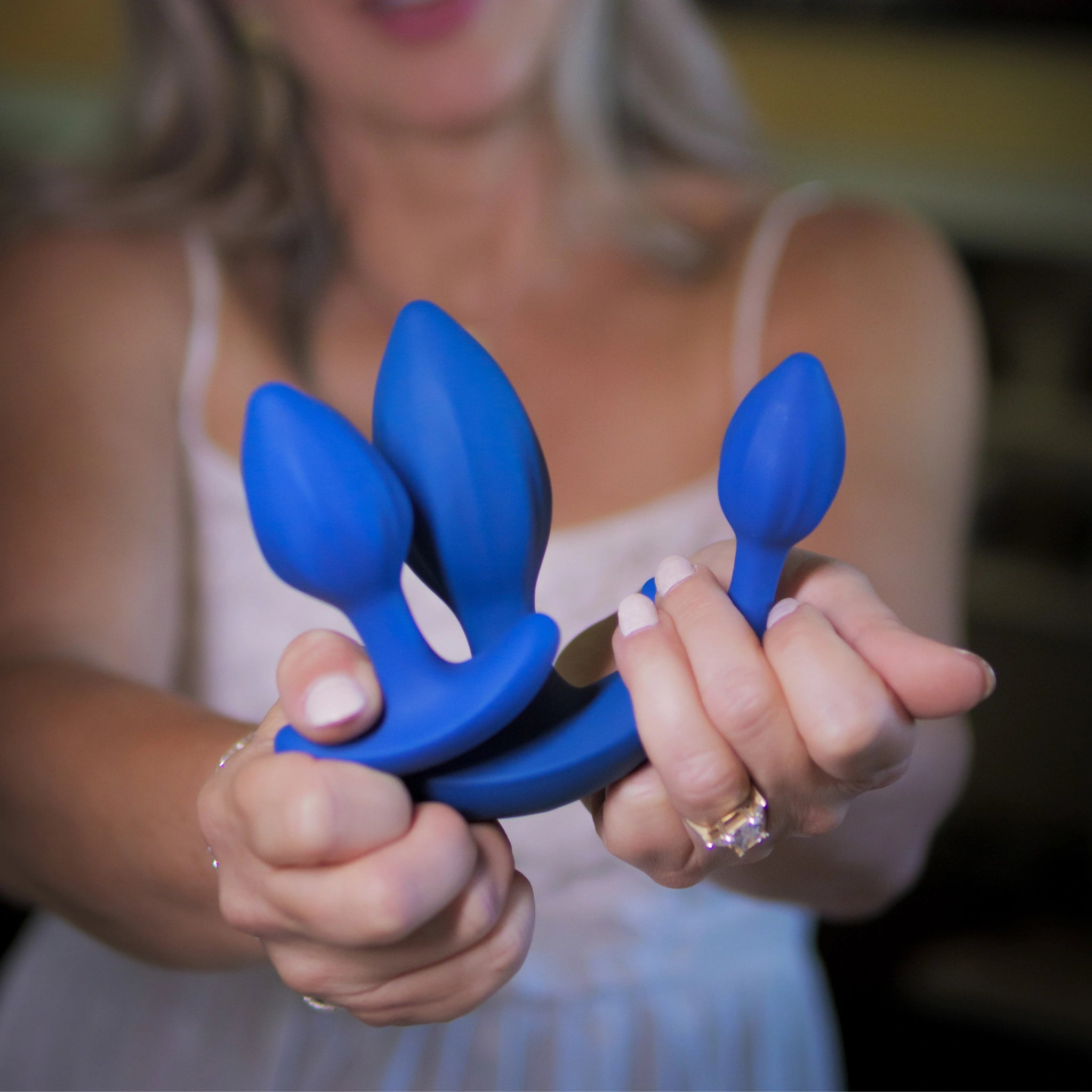 A woman in a white dress holds three blue silicone anal plugs of different sizes, displaying their shape and flexibility.