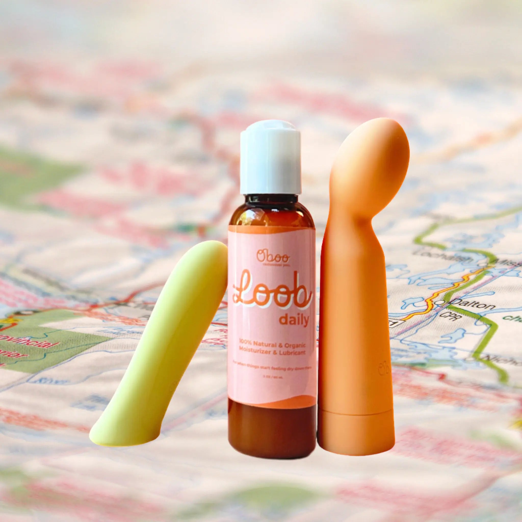 A bottle of Oboo Loob Daily lubricant is placed between two colorful massagers on a soft, blurred background of a map.