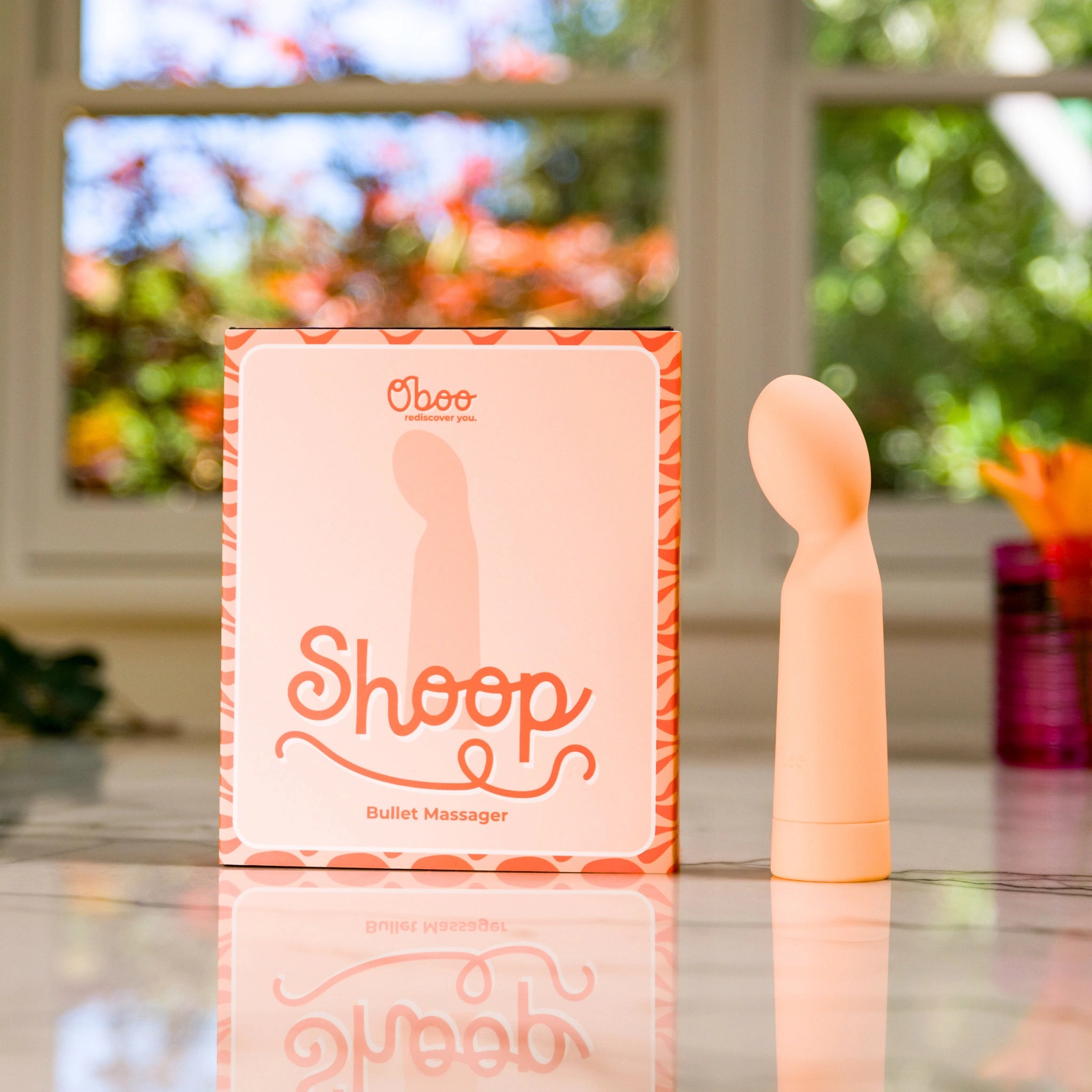 Shoop bullet massager displayed next to its branded packaging on a counter with colorful plants and a window in the background.