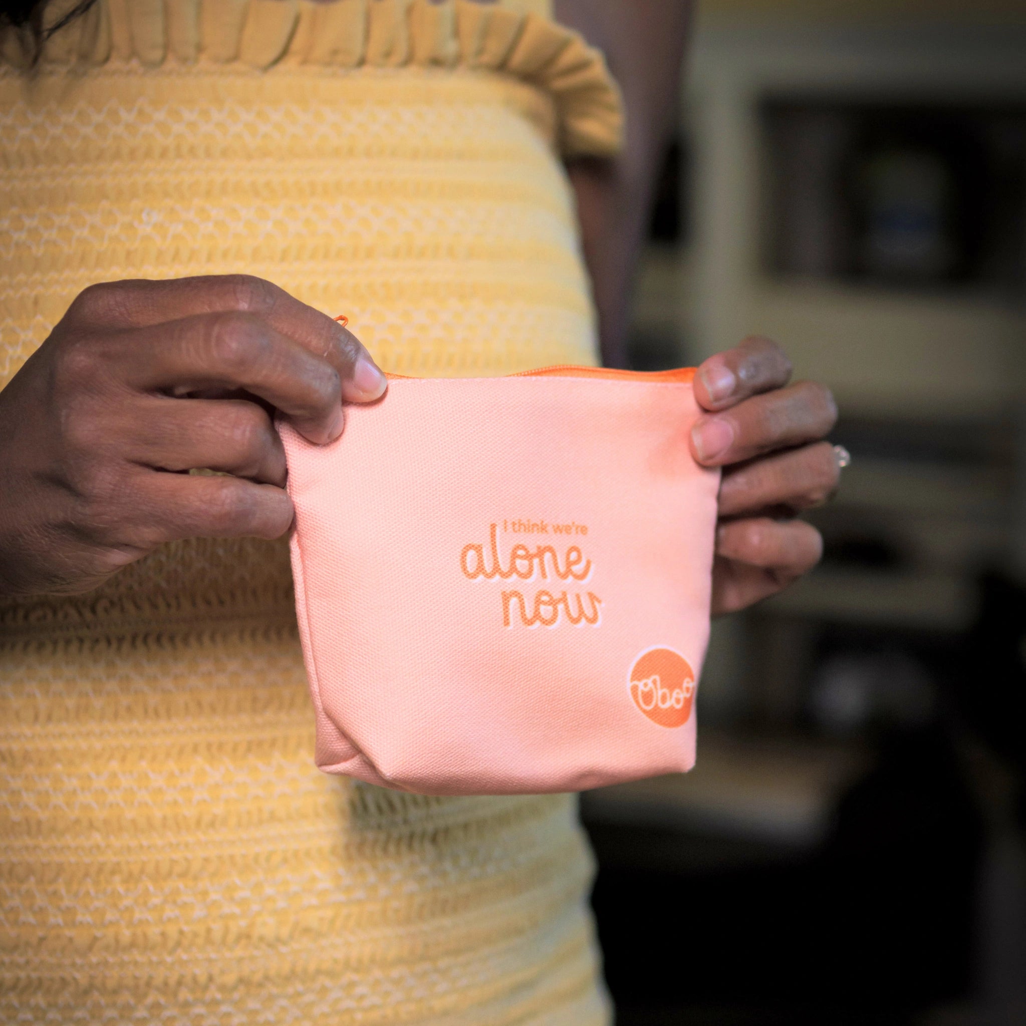 A peach-colored pouch with the words “I think we’re alone now” held by a woman wearing a yellow textured top.