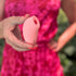 A woman in a pink dress holding a pink silicone stimulator in her hand, outdoors with greenery in the background.