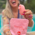 A smiling woman by a pool holding the pink stimulator above a matching pink pouch with playful embroidered text.
