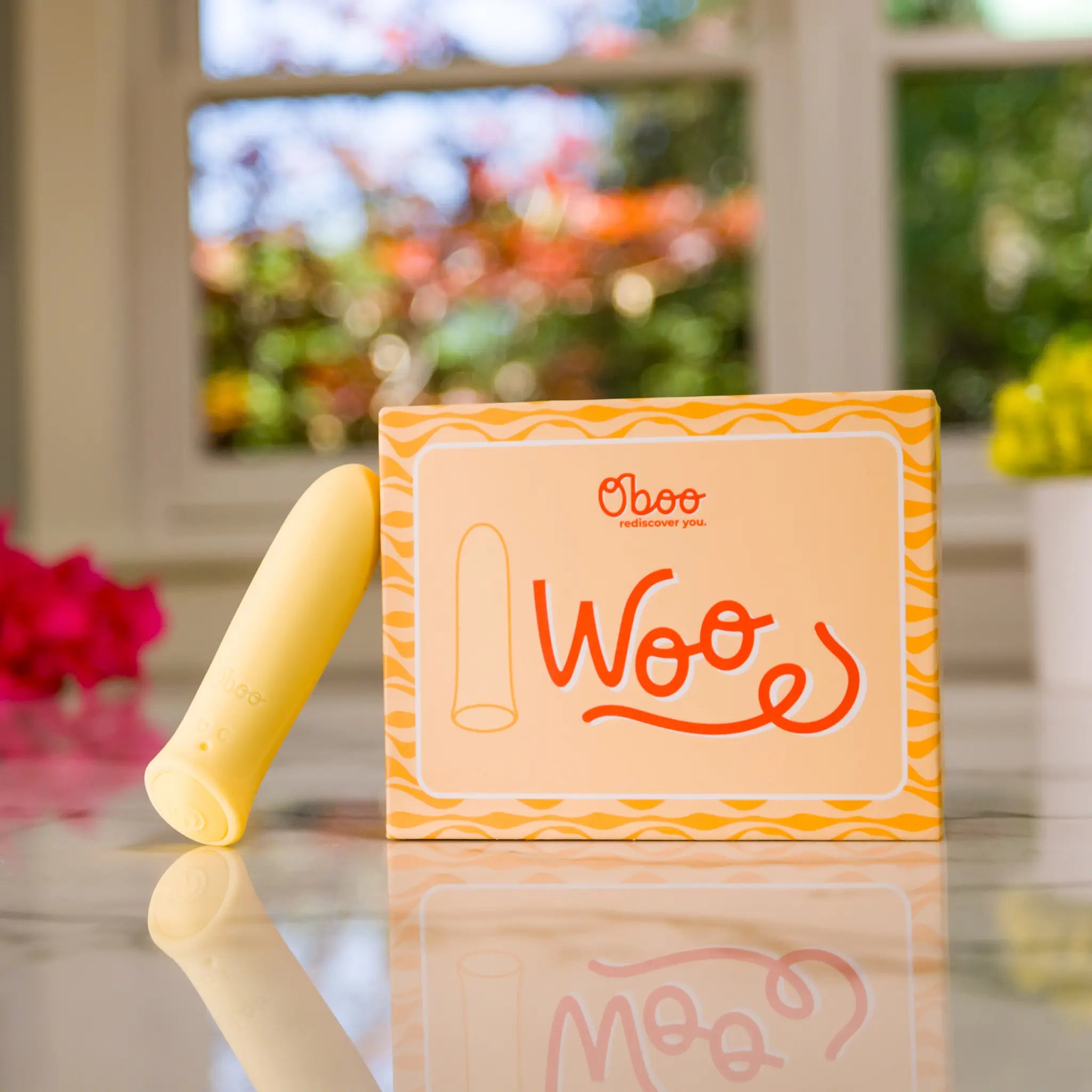 A yellow Woo massager stands beside its matching Oboo product box on a reflective surface, with soft natural light in background.