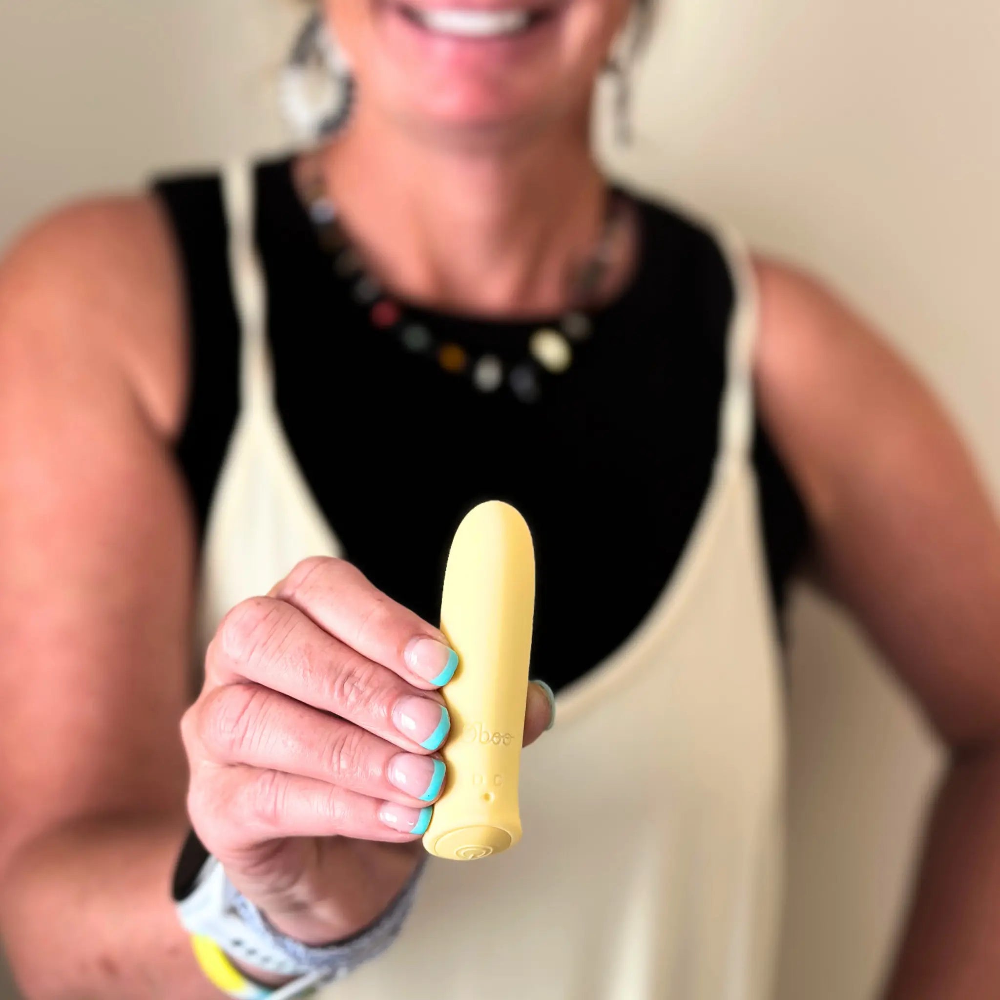 A smiling woman in a cream dress holds a yellow Woo massager close to the camera, highlighting its compact, rounded design.