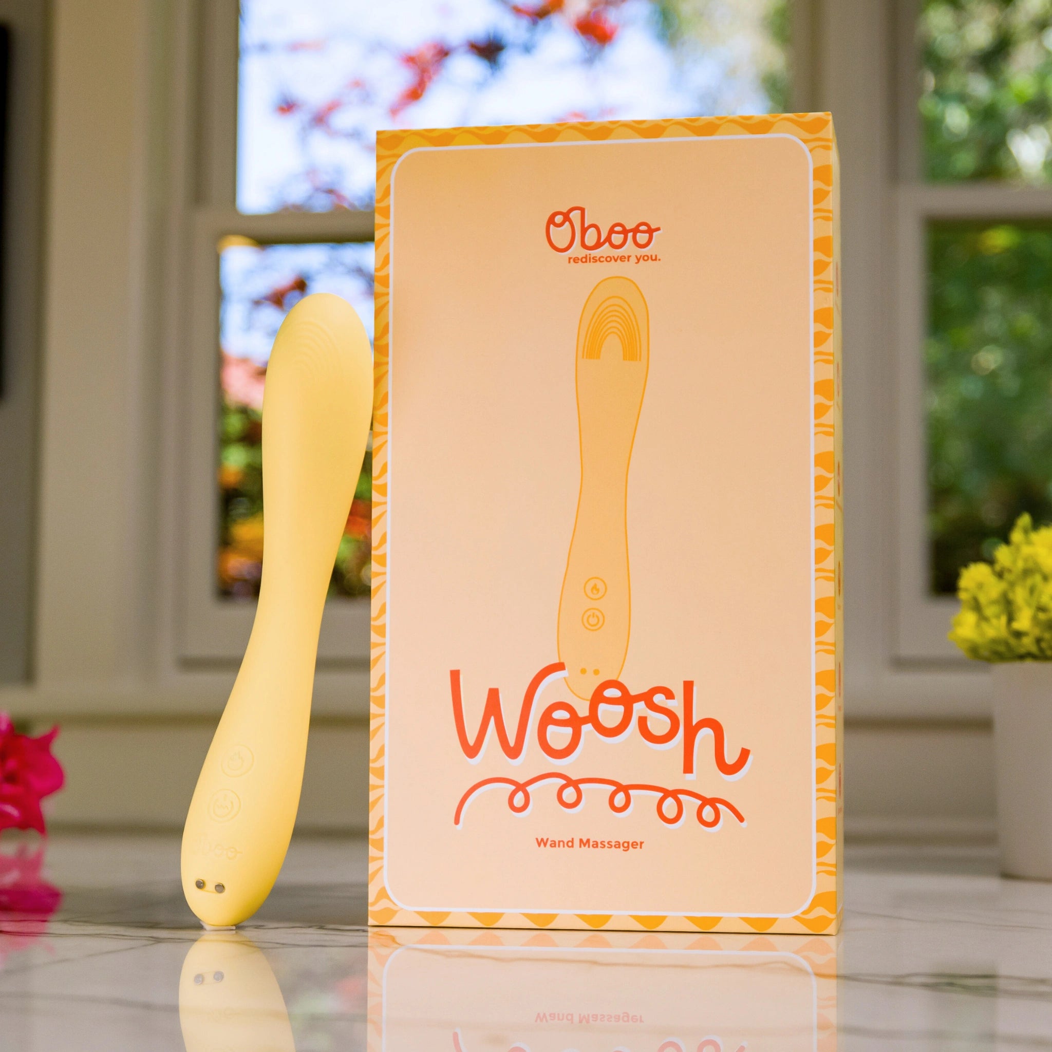 Yellow wand massager standing next to its bright packaging that reads “Woosh” by Oboo, placed on a sunny windowsill.