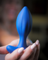 A woman with light pink nails holds a large blue silicone anal plug close to the camera, showcasing its smooth, tapered shape.