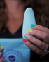 A woman with colorful rainbow nails holds a light blue flexible massager, partially visible against her chest and a blue pouch.