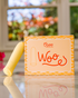A yellow Woo massager stands beside its matching Oboo product box on a reflective surface, with soft natural light in background.