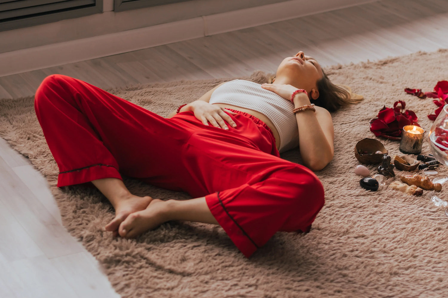 A woman in red pants lies on a soft rug with her hand on her stomach, surrounded by candles, crystals, and rose petals.