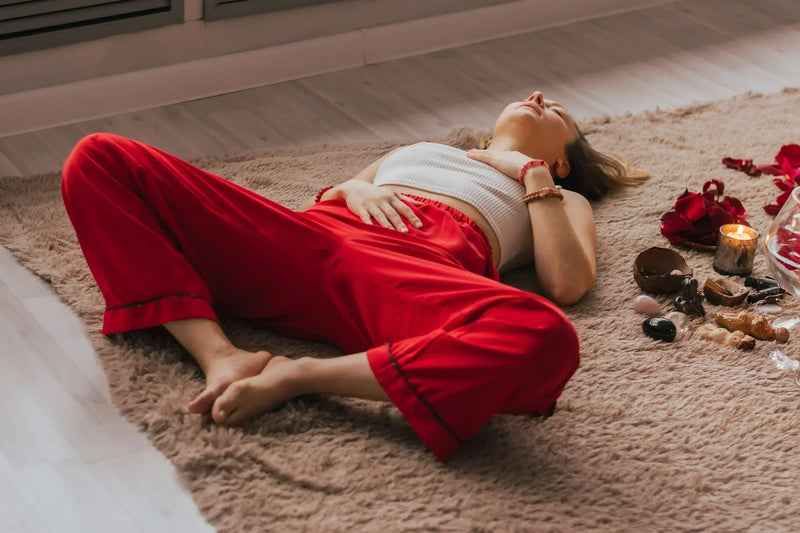 A woman in red pants lies on a soft rug with her hand on her stomach, surrounded by candles, crystals, and rose petals.