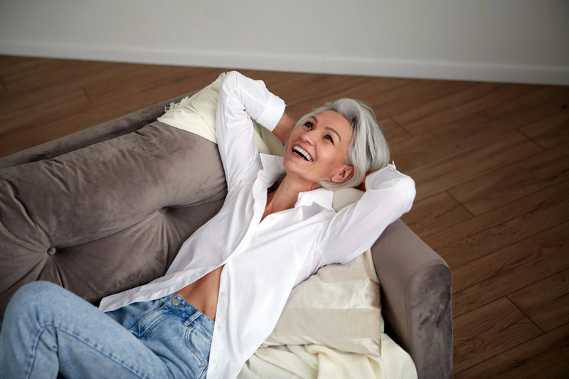 Middle-aged woman with gray hair smiling and relaxing on a soft couch, wearing a white shirt and jeans in a cozy room.