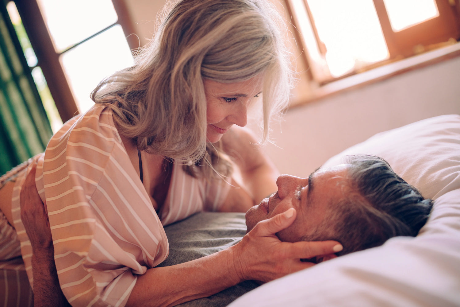 Older couple shares an intimate, loving moment in bed, with the woman smiling warmly as she gazes into her partner’s eyes.