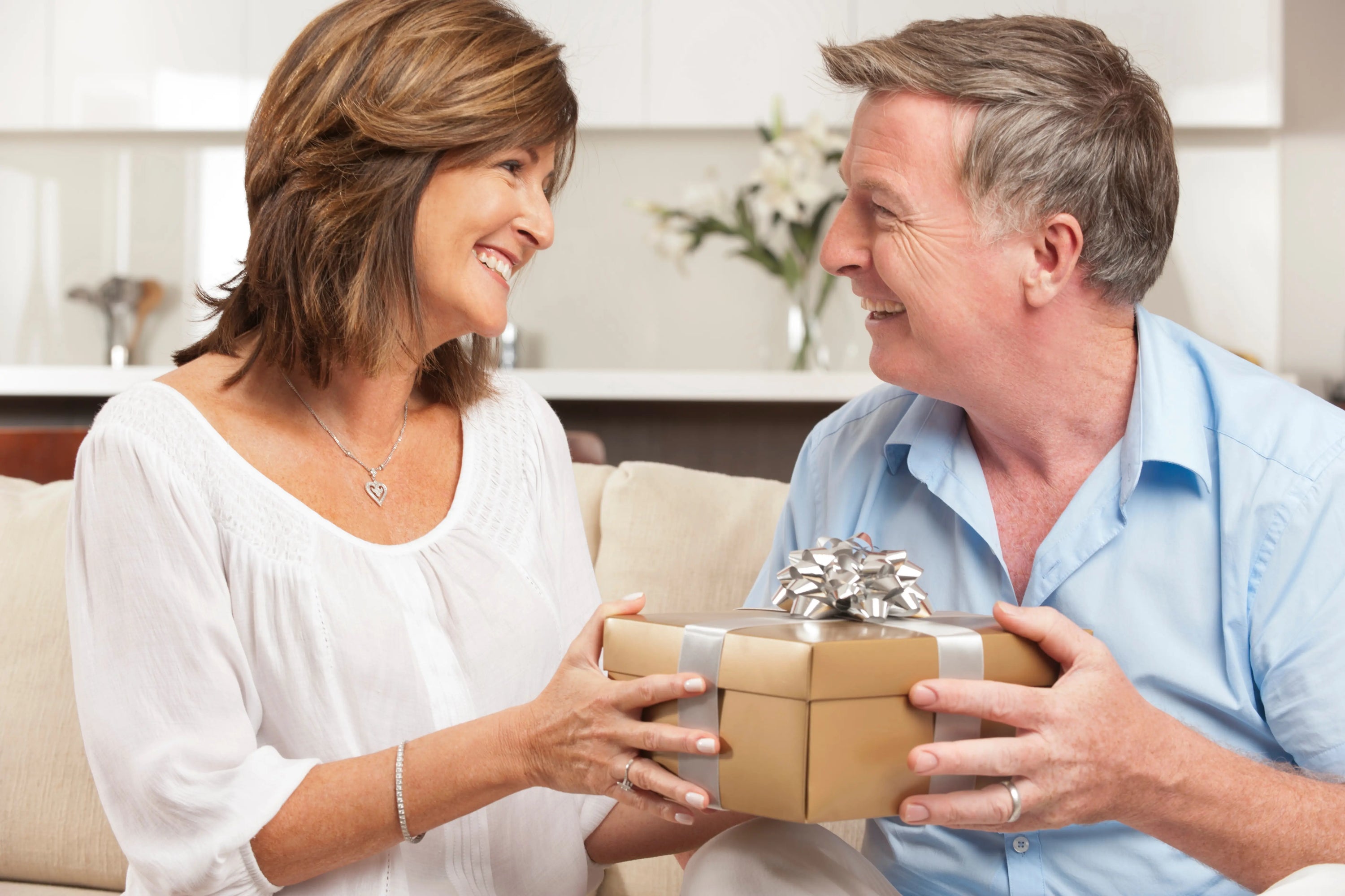 A smiling middle-aged couple sits on a couch, exchanging a gold-wrapped gift with a silver ribbon and bow.