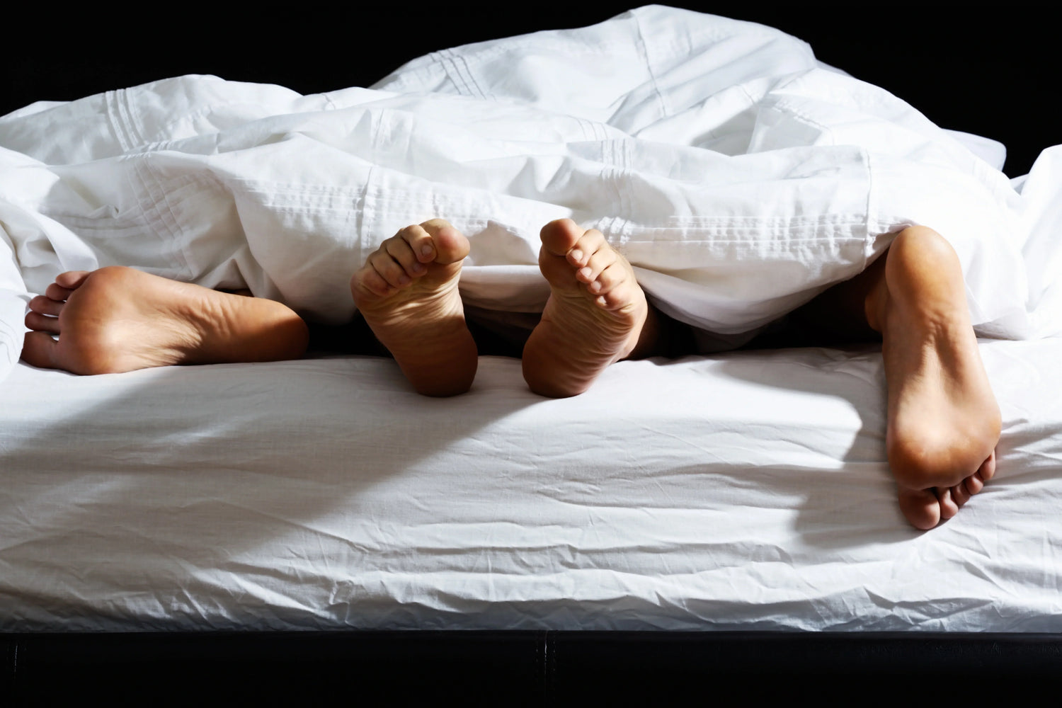 Three pairs of feet peek out from under a white blanket on a bed, suggesting intimacy or playful closeness among people.