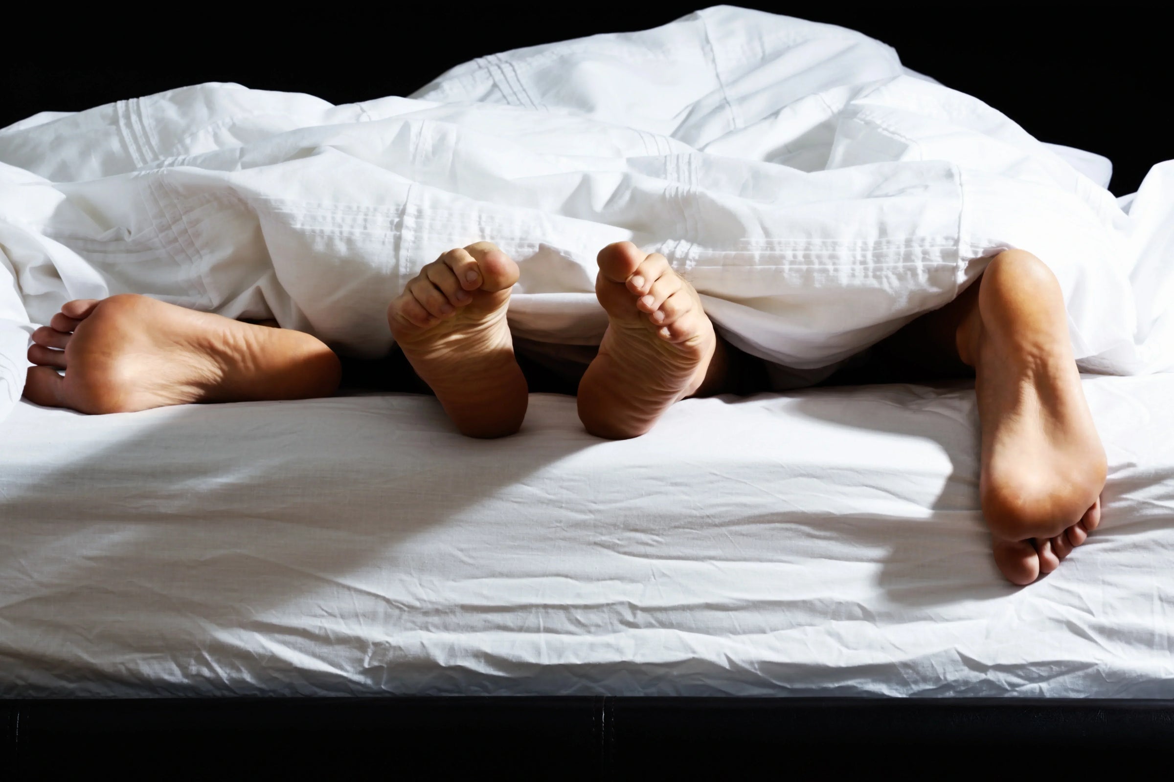 Three pairs of feet peek out from under a white blanket on a bed, suggesting intimacy or playful closeness among people.