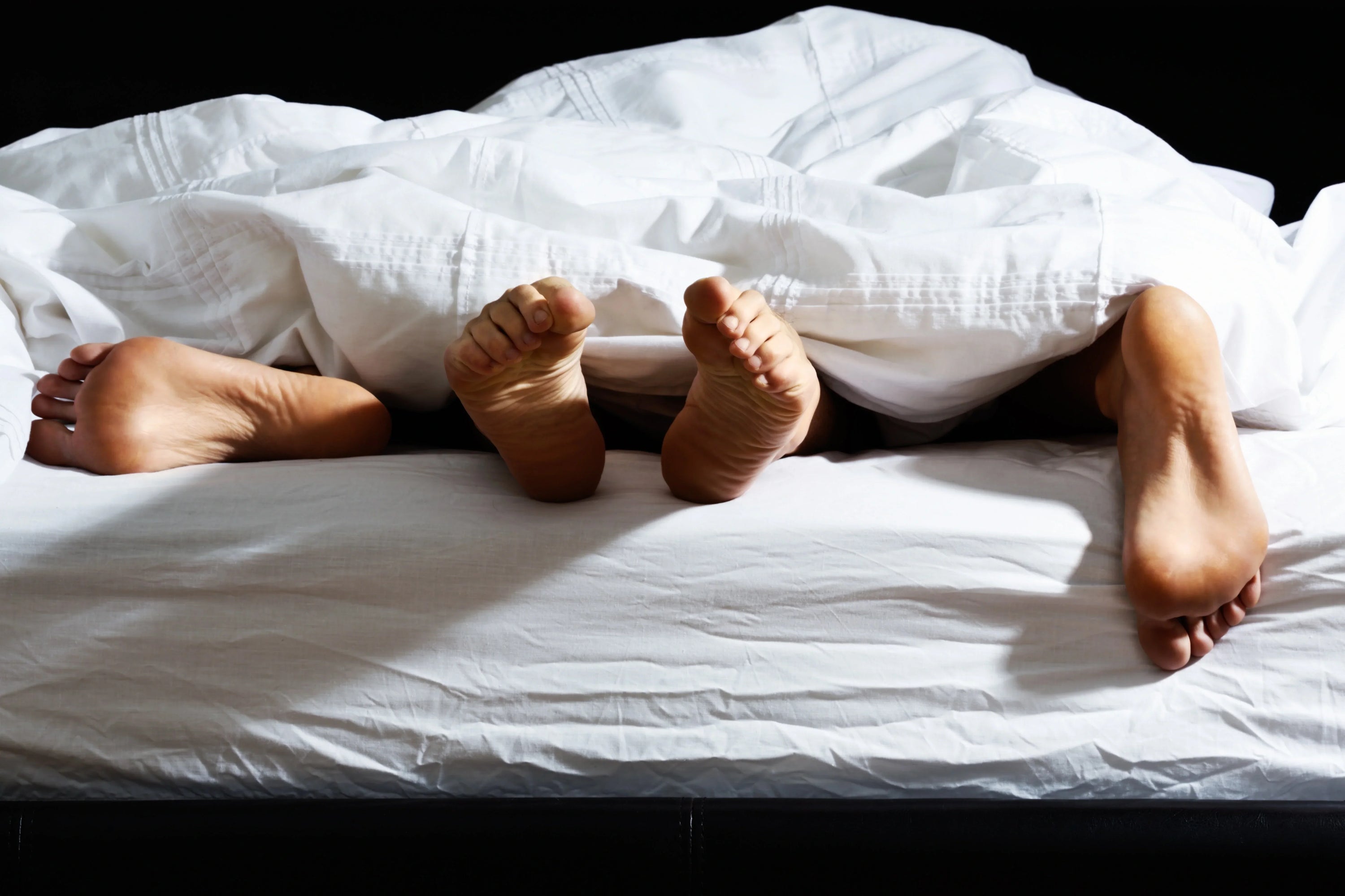 Three pairs of feet peek out from under a white blanket on a bed, suggesting intimacy or playful closeness among people.