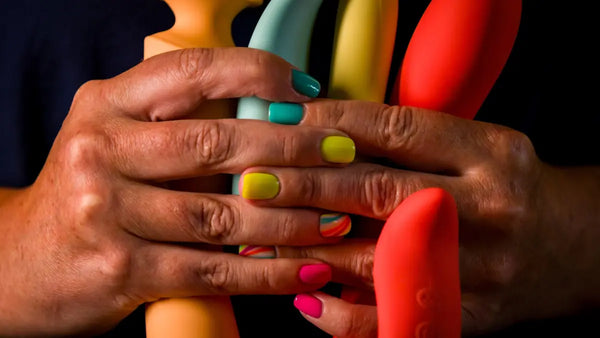 Close-up of multiple hands with colorful nail polish stacked together, with bright red and green nails and a blurred background of colorful objects.