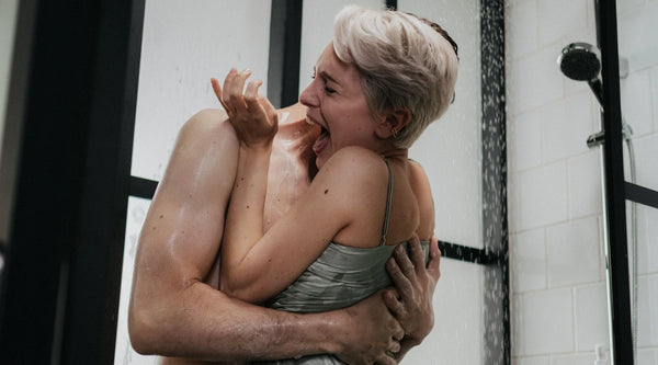 Person with short gray hair in a wet green clothes playfully splashes water with hands while standing in a shower stall, with tiled wall and shower fixture visible.