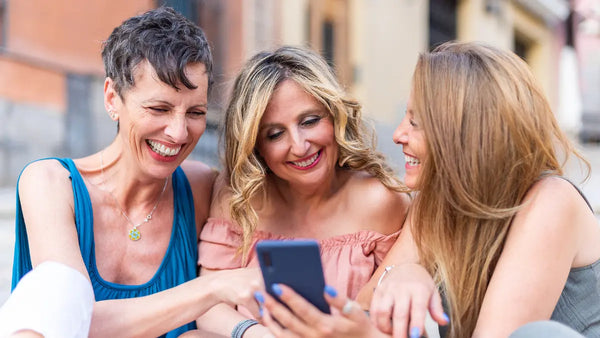 Three friends outdoors smiling and leaning in as one holds a smartphone, sharing a moment and looking at the screen.