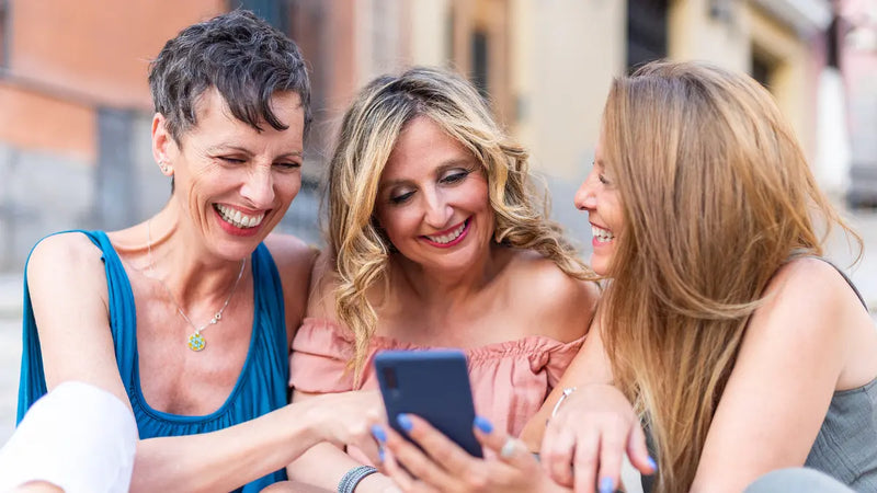 Three friends outdoors smiling and leaning in as one holds a smartphone, sharing a moment and looking at the screen.