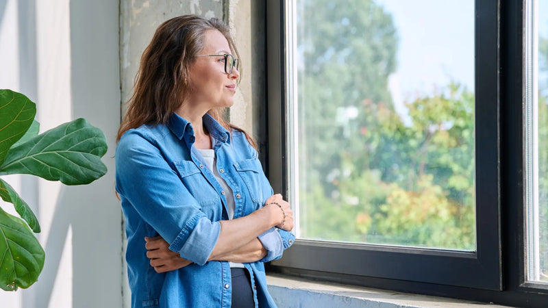 Middle-aged woman wearing glasses and denim shirt standing by window, arms crossed, thoughtfully looking outside with sunlight and greenery visible.