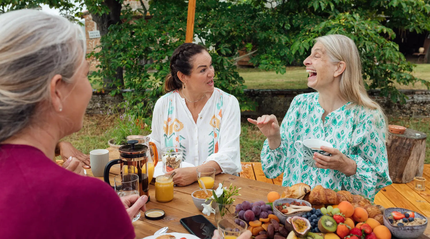 Three women laughing and talking around a wooden table laden with fruit, pastries, and drinks outdoors.
