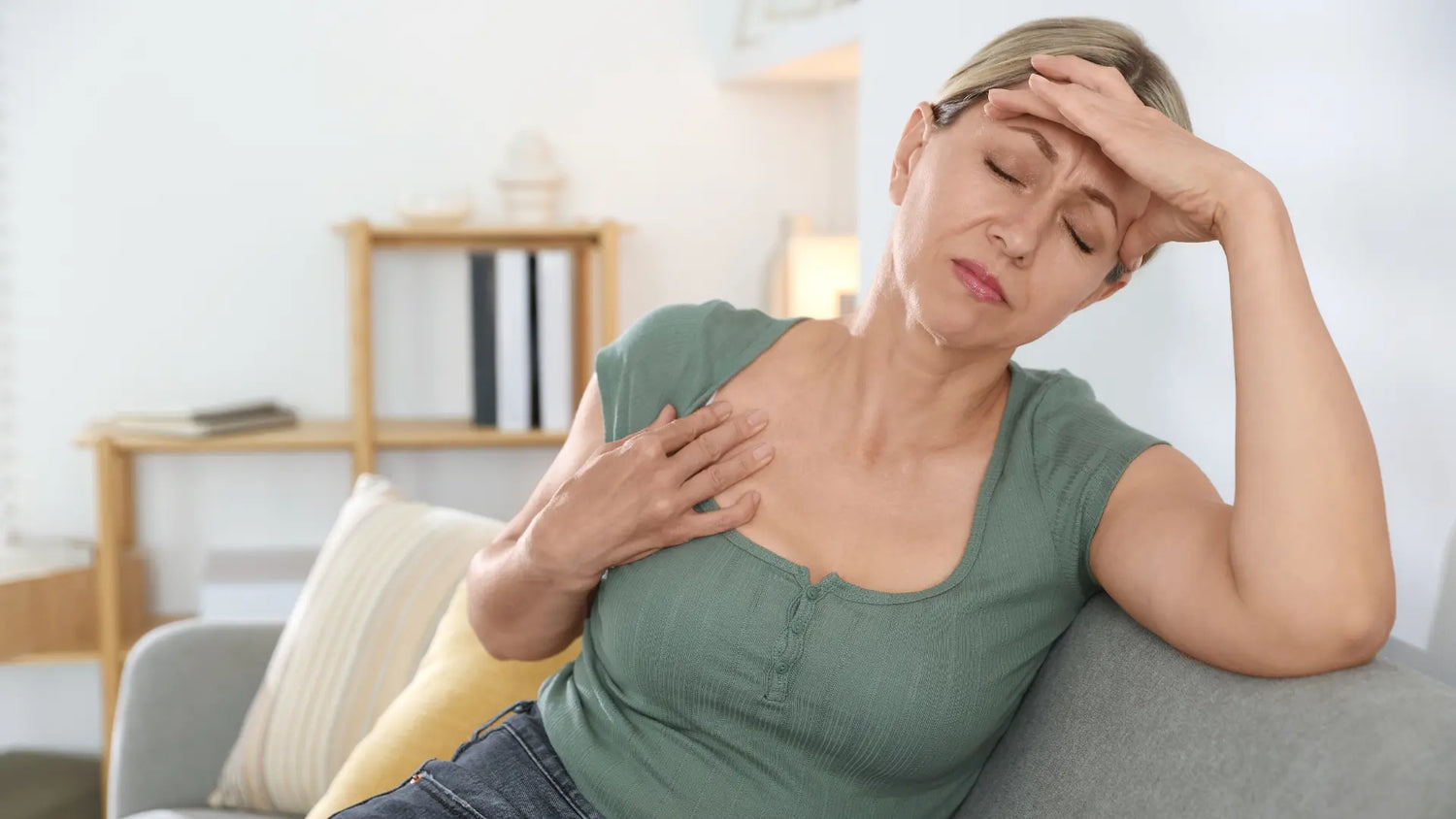 A midlife woman sits on a couch, hand on forehead and chest, looking uncomfortable during a hot flash.