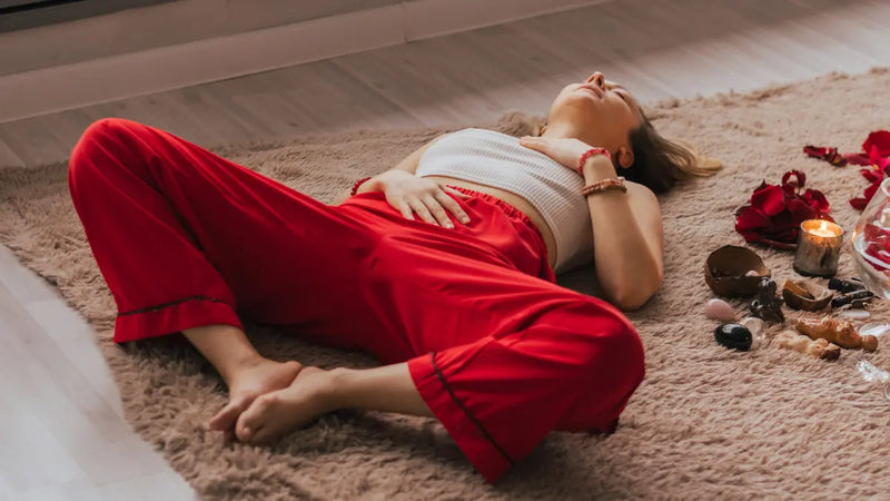 A person lies on a rug with legs in a butterfly pose, hands resting on their chest and abdomen. A collection of crystals, rose petals, and a lit candle are arranged to the side.