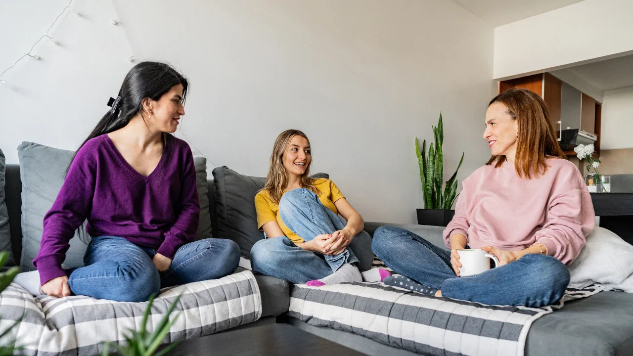 Three women sit together on a couch in a cozy living room, chatting and smiling while enjoying each other’s company.