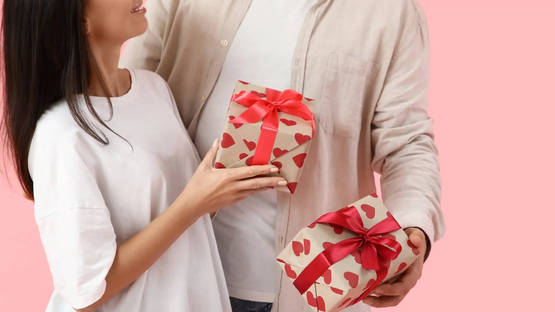 Couple exchanging heart wrapped gifts, smiling closely together against a pink background, celebrating love, intimacy, and Valentine’s Day moment joy.
