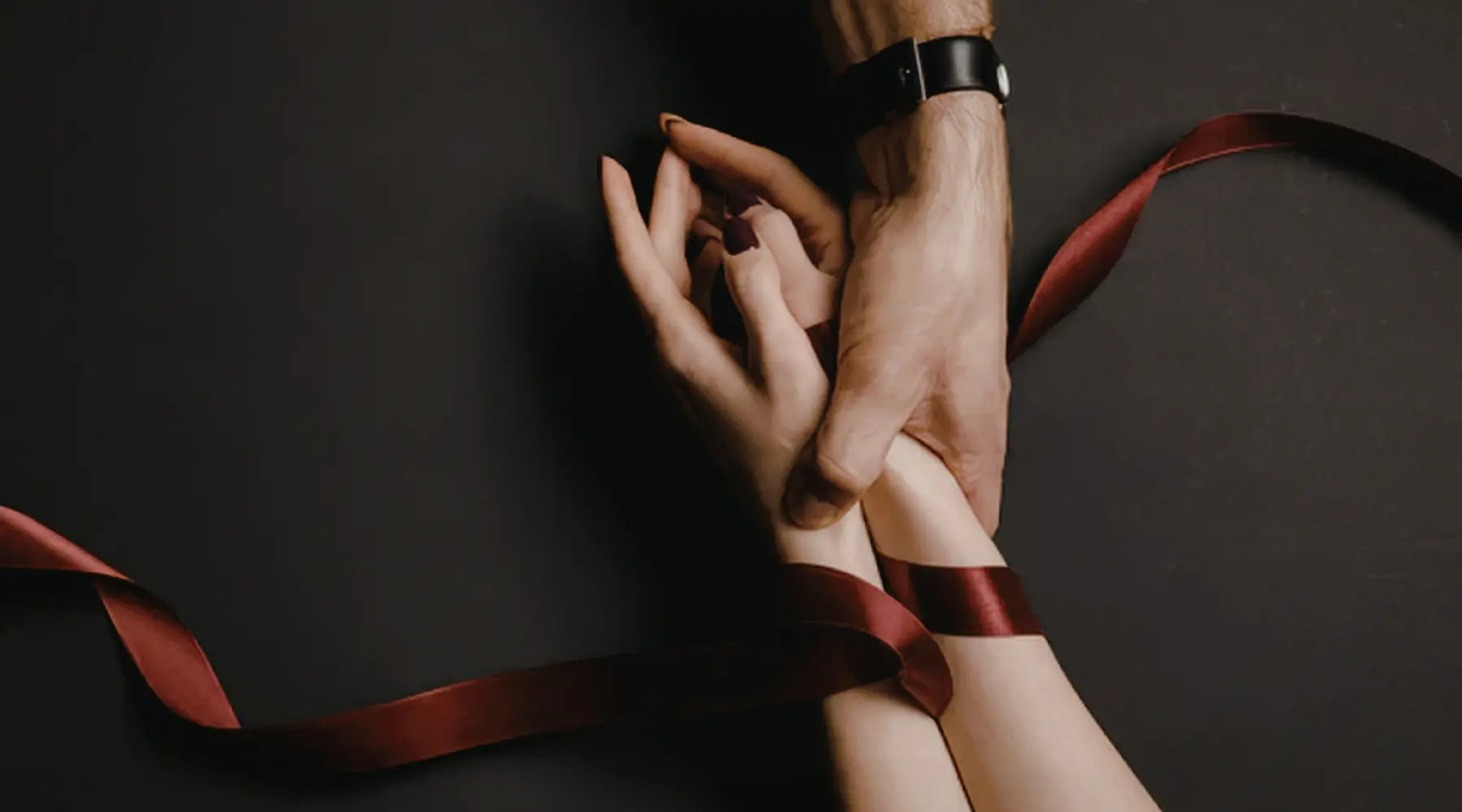 A close-up shot of hands intertwined, bound by a deep red ribbon against a dark background.