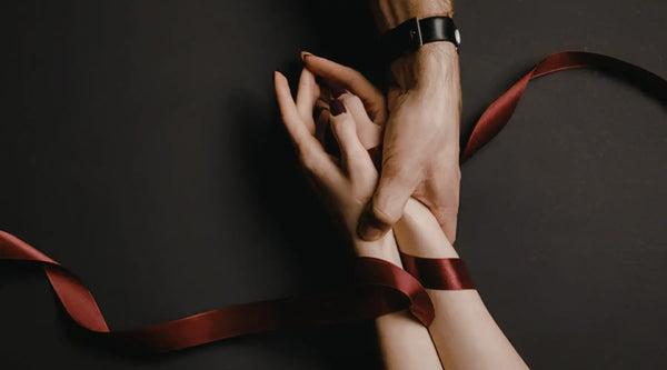 A close-up shot of hands intertwined, bound by a deep red ribbon against a dark background.
