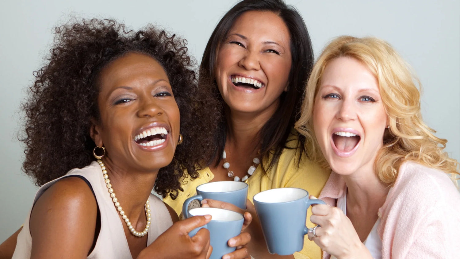 Three midlife women laughing together while holding coffee mugs, smiling closely in a studio setting, celebrating friendship, warmth, and connection.