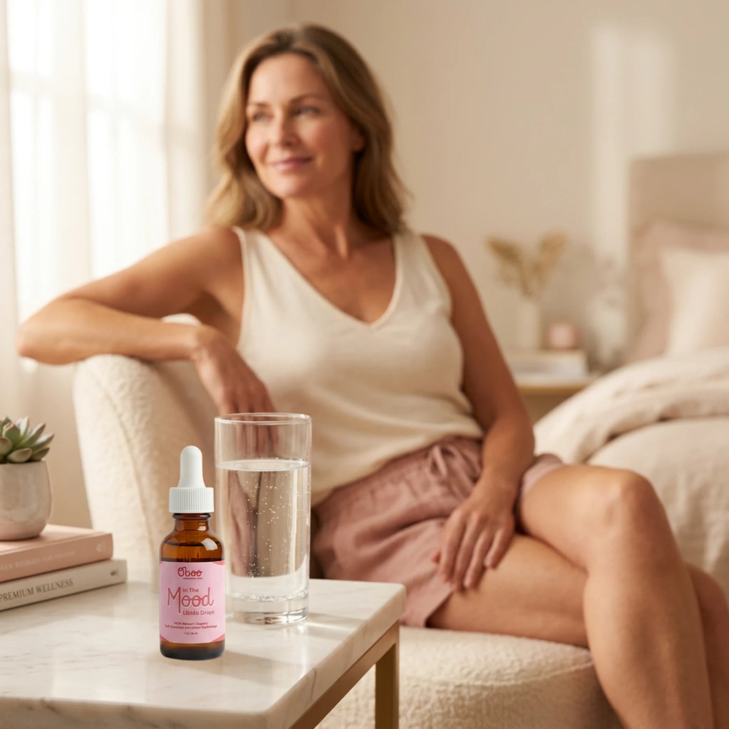 Smiling midlife woman in a cream tank top relaxing in her bedroom, In the Mood Drops and a glass of water on the side table beside her