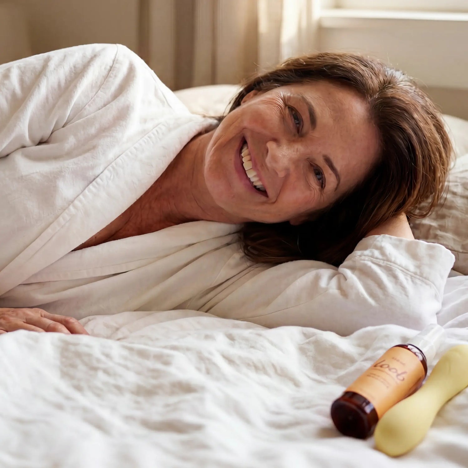Midlife woman laughing softly in white linens with morning light, a yellow Loob Arousal bottle beside her on the bed