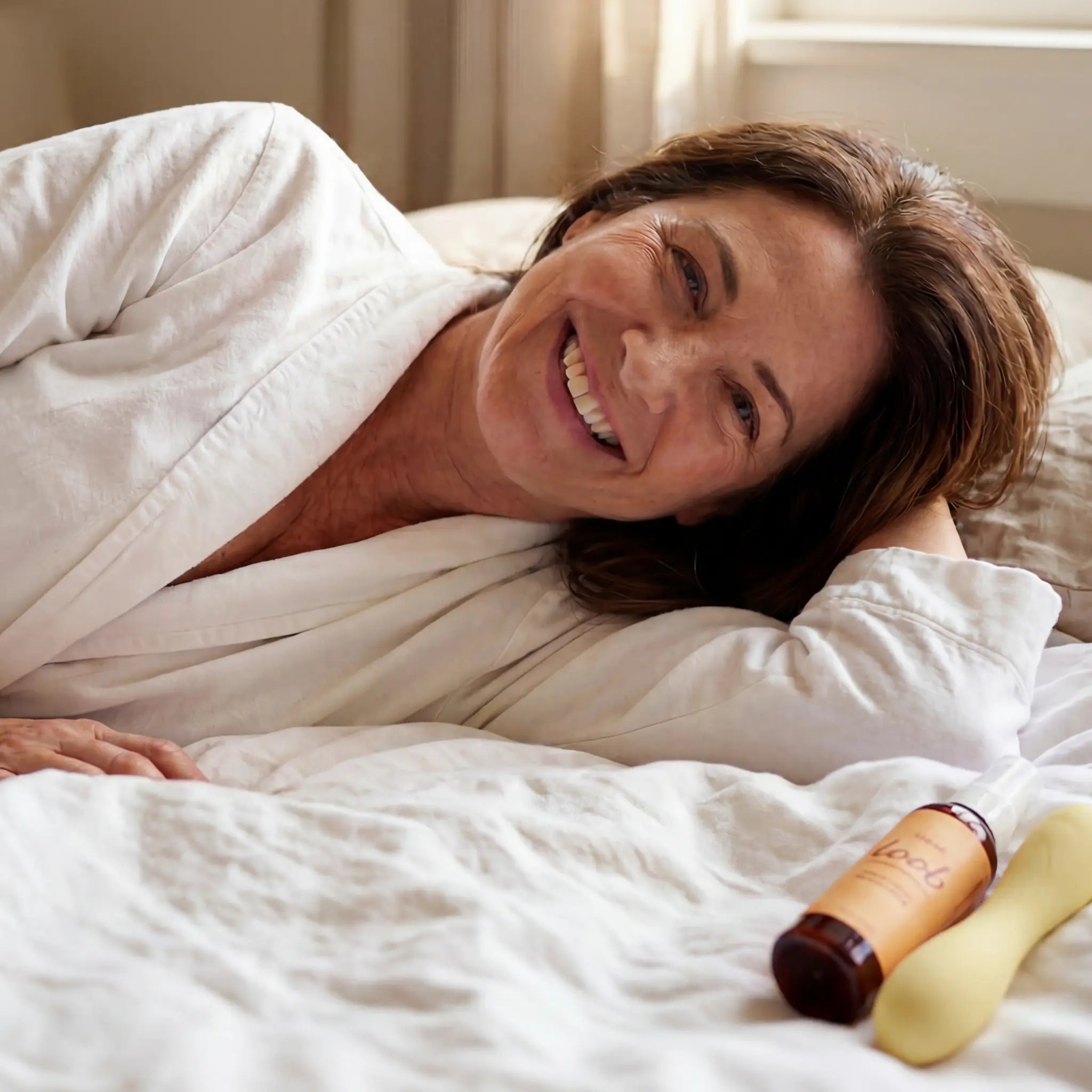 Midlife woman laughing softly in white linens with morning light, a yellow Loob Arousal bottle beside her on the bed