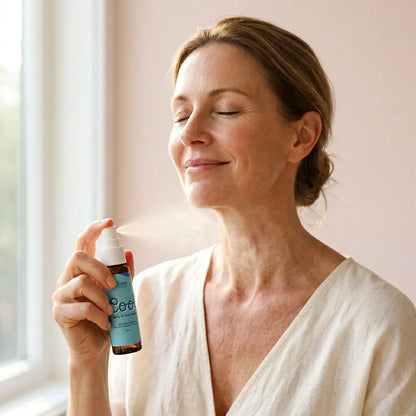 Smiling midlife woman in a cream linen robe spritzing Oboo Cool Spray on her neck, eyes closed, soft natural light from the window