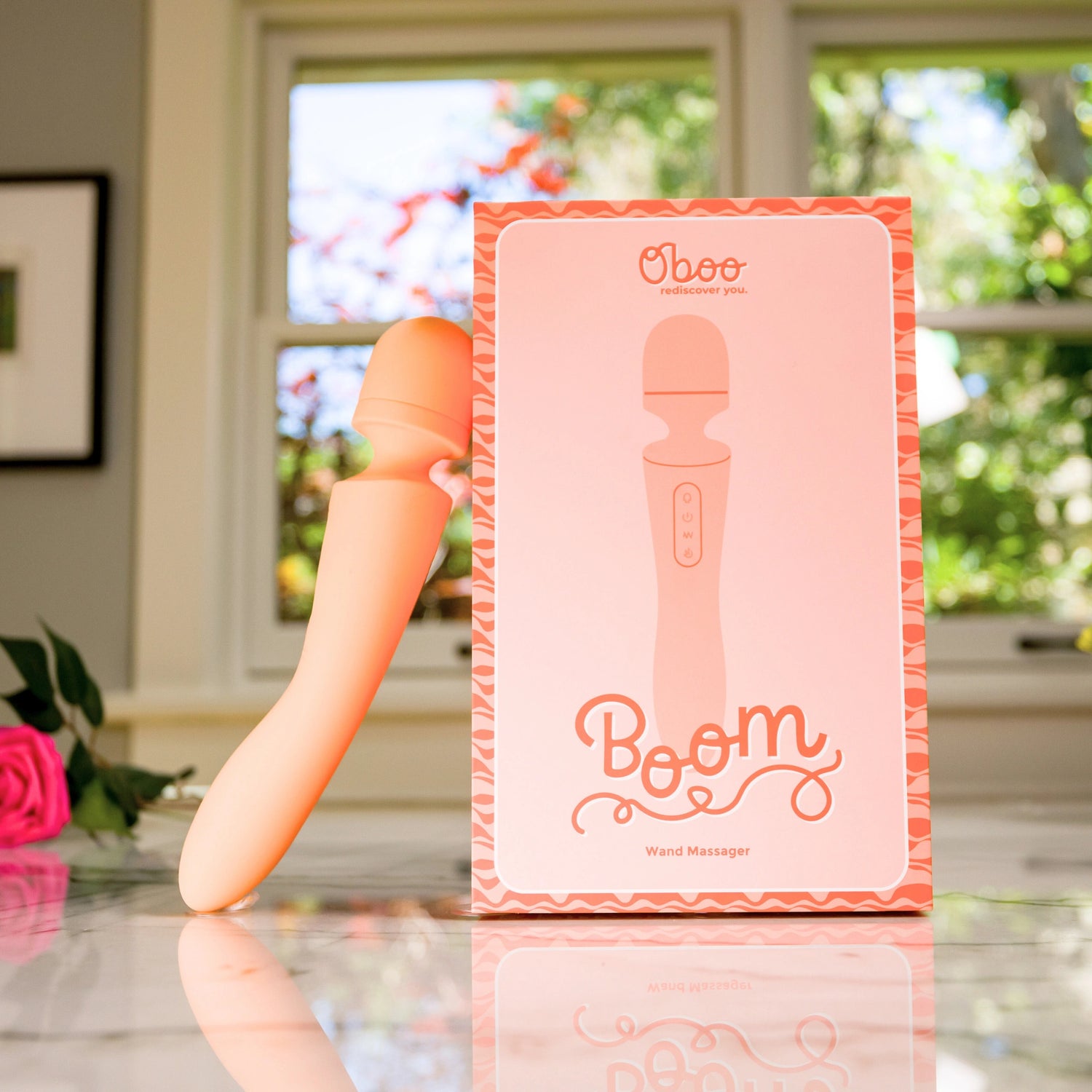Boom wand massager standing next to its pink product box with branding, placed on a marble countertop near a window.