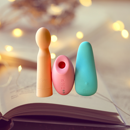 Three colorful Oboo massagers—peach, pink, and teal—stand upright on an open book with warm bokeh lights in the background.