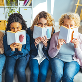 Three women sit side by side, playfully hiding their faces behind books that feature pixelated images of colorful Oboo massagers.