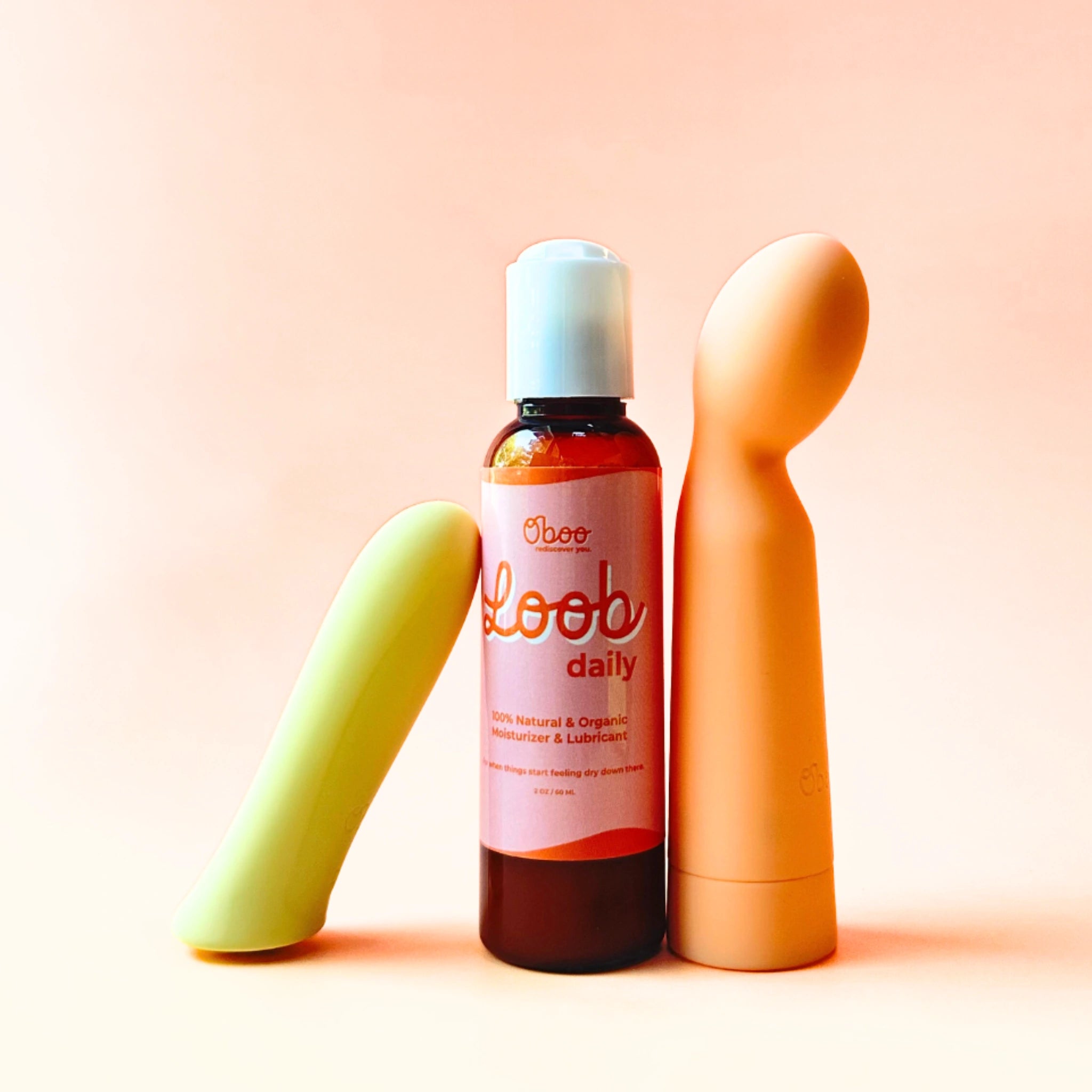 Loob Daily bottle stands upright between two colorful vibrators on a peach background, suggesting pleasure, comfort, and sensual self-care.