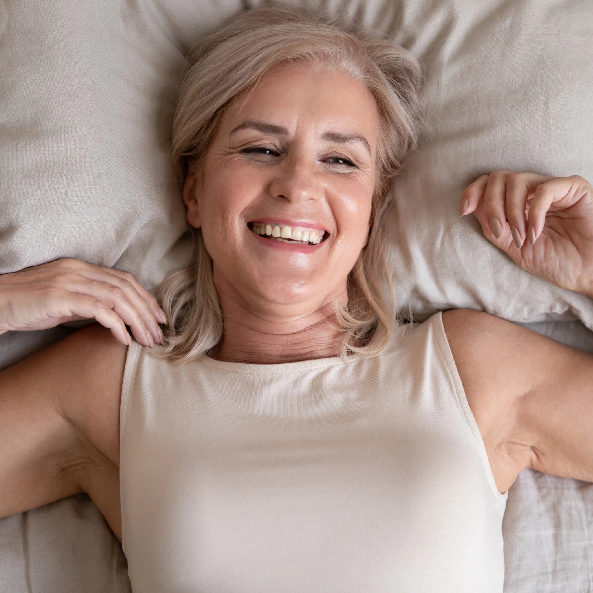 Smiling midlife woman lying comfortably on a bed with neutral bedding, suggesting confidence, relaxation, and contentment in her body.