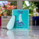 A light blue flexible massager stands next to its matching Oboo Oooh product box on a reflective surface near a window.