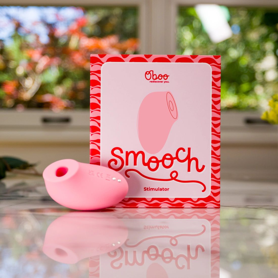 A pink clitoral stimulator placed in front of its product box labeled &quot;Smooch&quot; with a bright, colorful background.